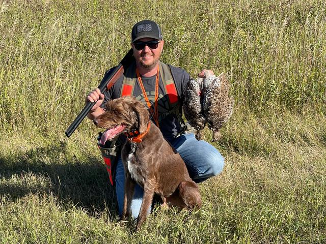 Dave kneeling in tall grass with his brown dog and sharp-tailed grouse